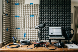 Mesa de home studio com microfone e interface, mostrando parede de fundo dividida entre caixas de ovo com fita azul e painel de espuma acústica piramidal preta.